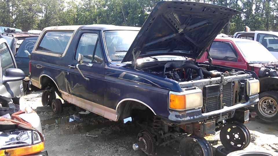 Blue Ford Bronco with hood open, missing wheels, in a junkyard. Another red vehicle is behind it