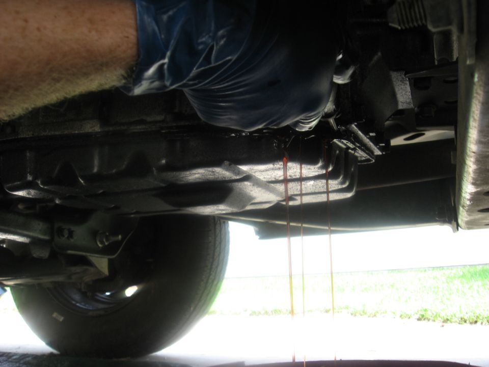 Transmission fluid draining from pan onto the ground. A gloved hand is visible near the pan
