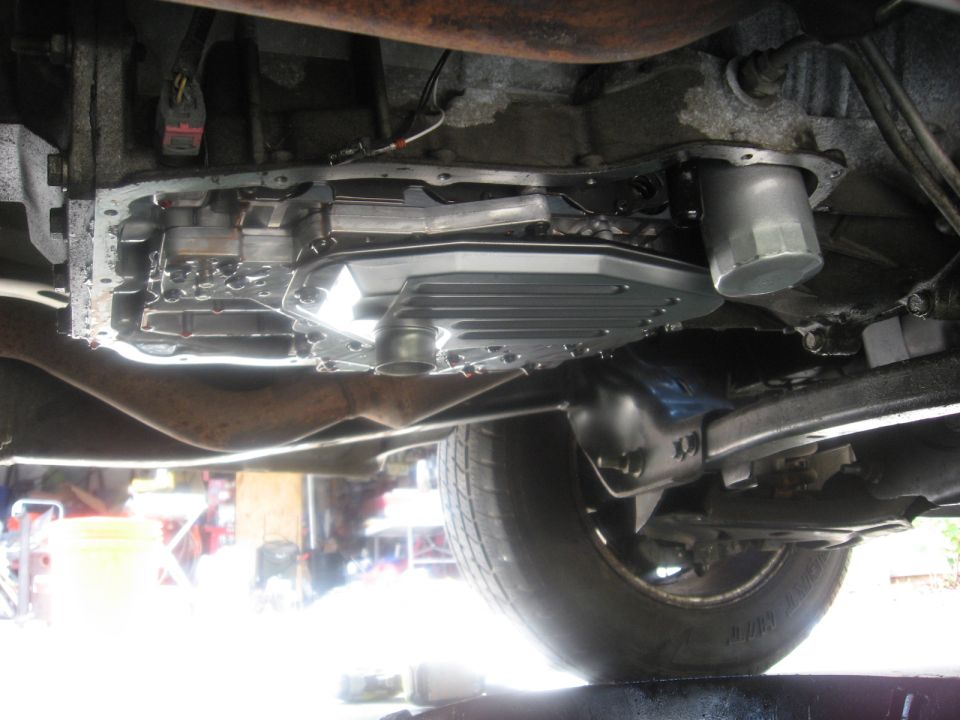 Underside view of a transmission pan and filter assembly with exhaust pipe and suspension components visible