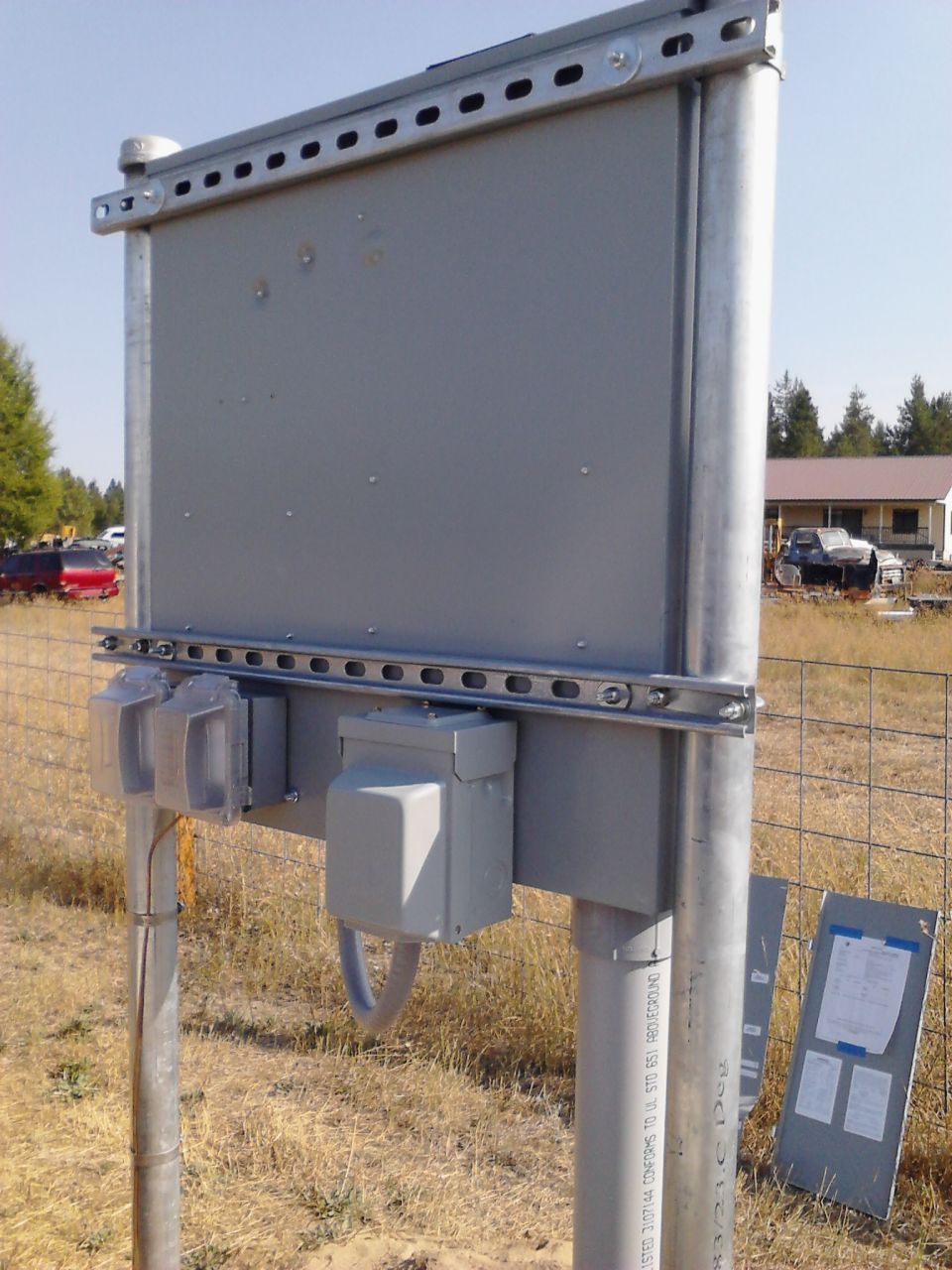 Gray electrical box mounted on metal poles, with a fence and some vehicles in the background