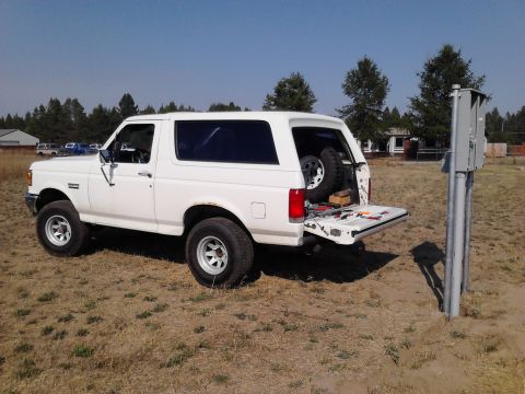 1989 Ford Bronco - A white Ford Bronco is parked outdoors with its tailgate open, revealing a spare tire and tools.