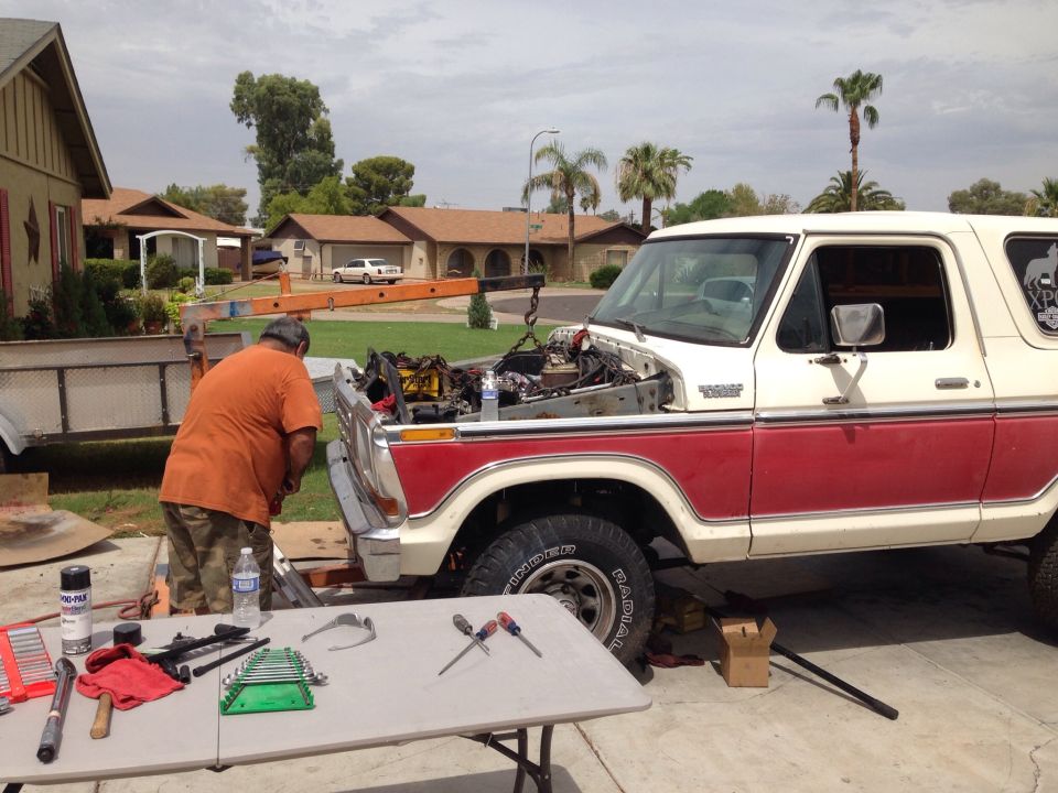 Man working on the engine of a red and white 1978 Ford Bronco, with tools laid out on a table