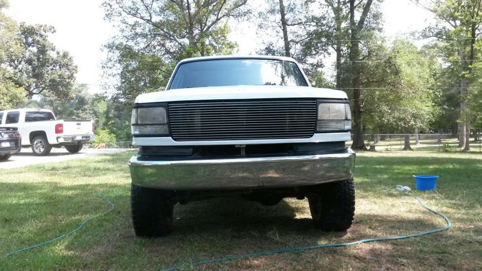 Front view of a white Ford F-Series truck with a chrome bumper and aftermarket grille. Another white pickup truck is parked to the left