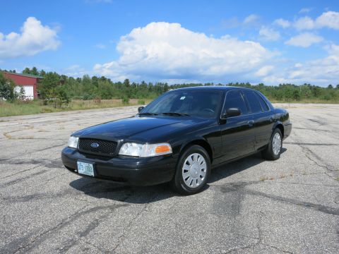 2001 Ford Crown Victoria - A black 2001 Ford Crown Victoria sedan is parked on a cracked asphalt lot under a blue sky with scattered clouds.