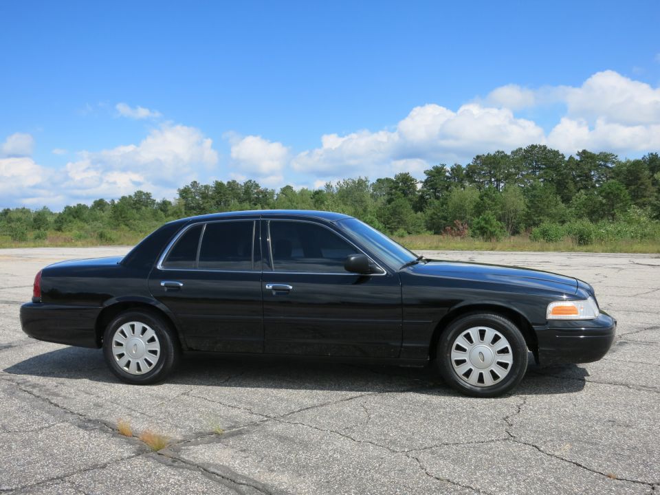 Black 2001 Ford Crown Victoria parked on asphalt with trees and blue sky in background