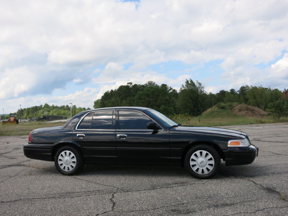 Black 2001 Ford Crown Victoria parked on asphalt with trees and cloudy sky in background