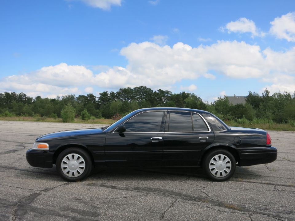 Black 2001 Ford Crown Victoria sedan parked on asphalt lot with trees and blue sky in background