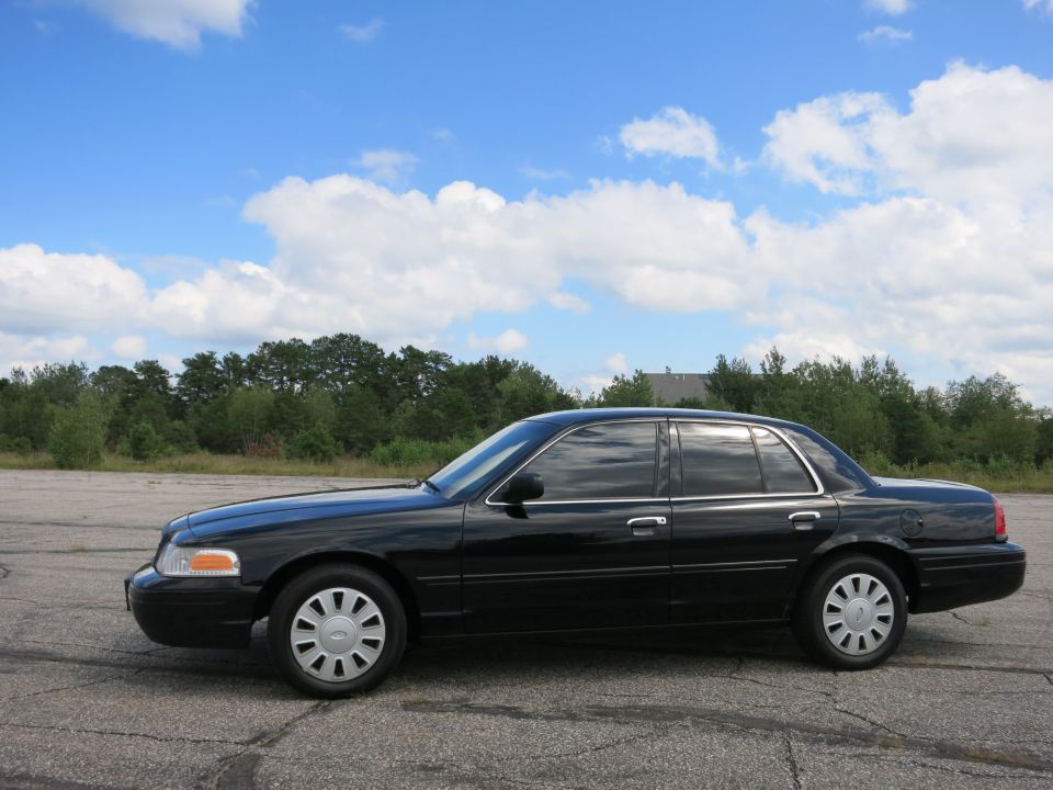 Black 2001 Ford Crown Victoria parked on asphalt with trees and blue sky in background