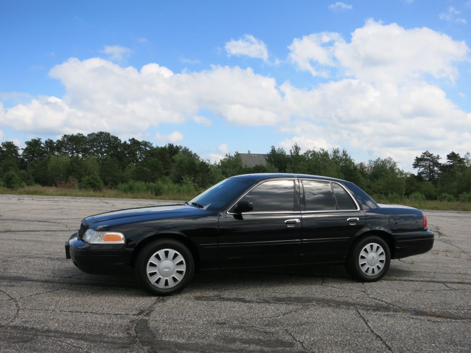 Black 2001 Ford Crown Victoria sedan, side view, parked on asphalt with trees and sky in background