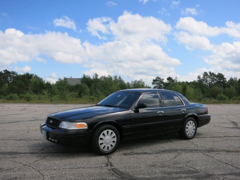 2001 Ford Crown Victoria - A black 2001 Ford Crown Victoria sedan is parked on an asphalt lot under a partly cloudy sky.