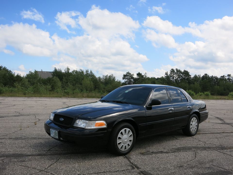 Black 2001 Ford Crown Victoria parked on asphalt lot with trees and sky in background