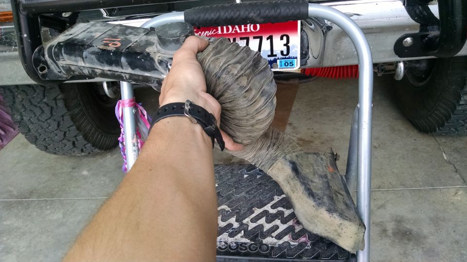 Hand holding a flexible air intake hose near a vehicle's rear bumper and tire. Idaho license plate visible