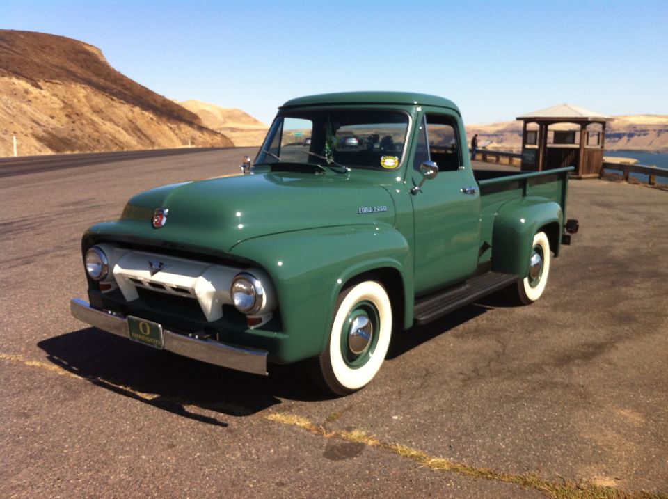 Green 1955 Ford F-100 pickup truck parked on a road with a view of hills and water