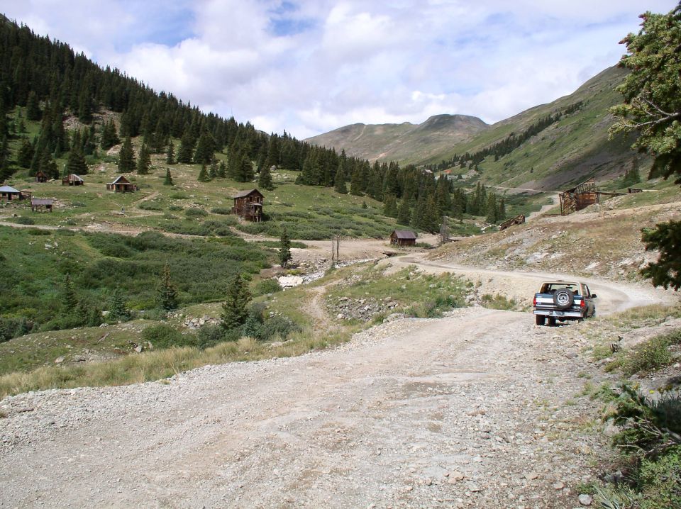 SUV parked on a dirt mountain road above a valley with old wooden cabins and pine slopes