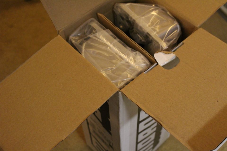 Open cardboard box containing two plastic-wrapped metal parts cushioned inside