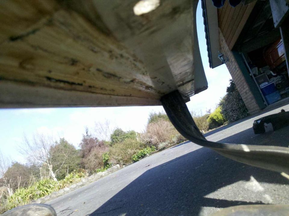 Underside view of a vehicle's fender and tire, showing dirt and wear. A garage is visible in the background