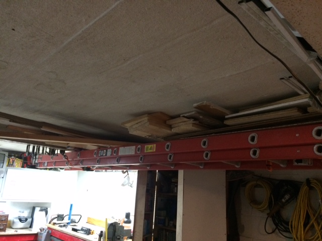 Red extension ladders are mounted overhead, holding lumber and other materials. A white cabinet and tools are visible below