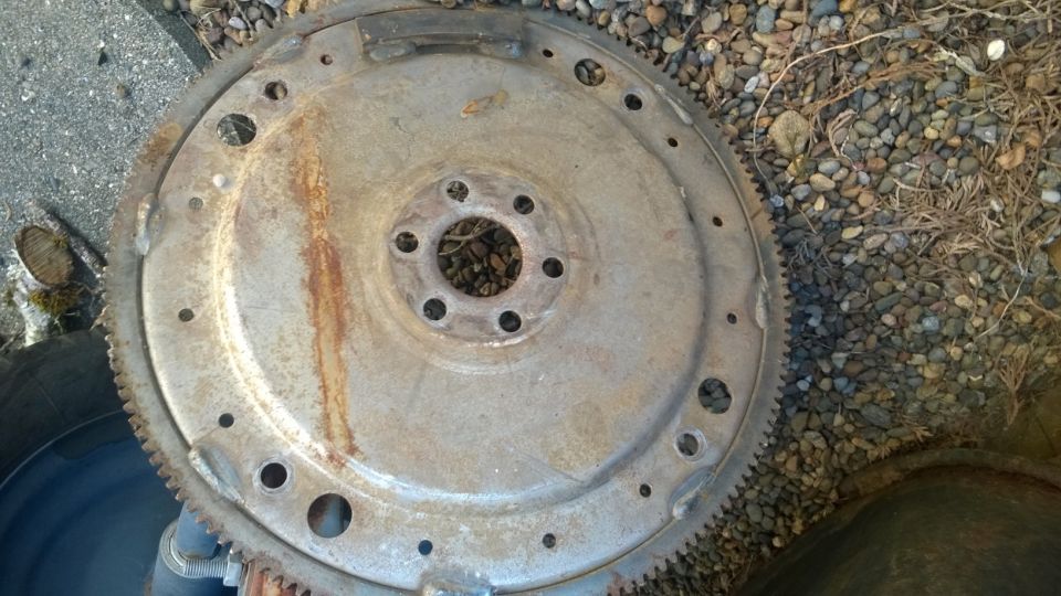Rusty metal flywheel with a ring of teeth and bolt holes, lying on gravel