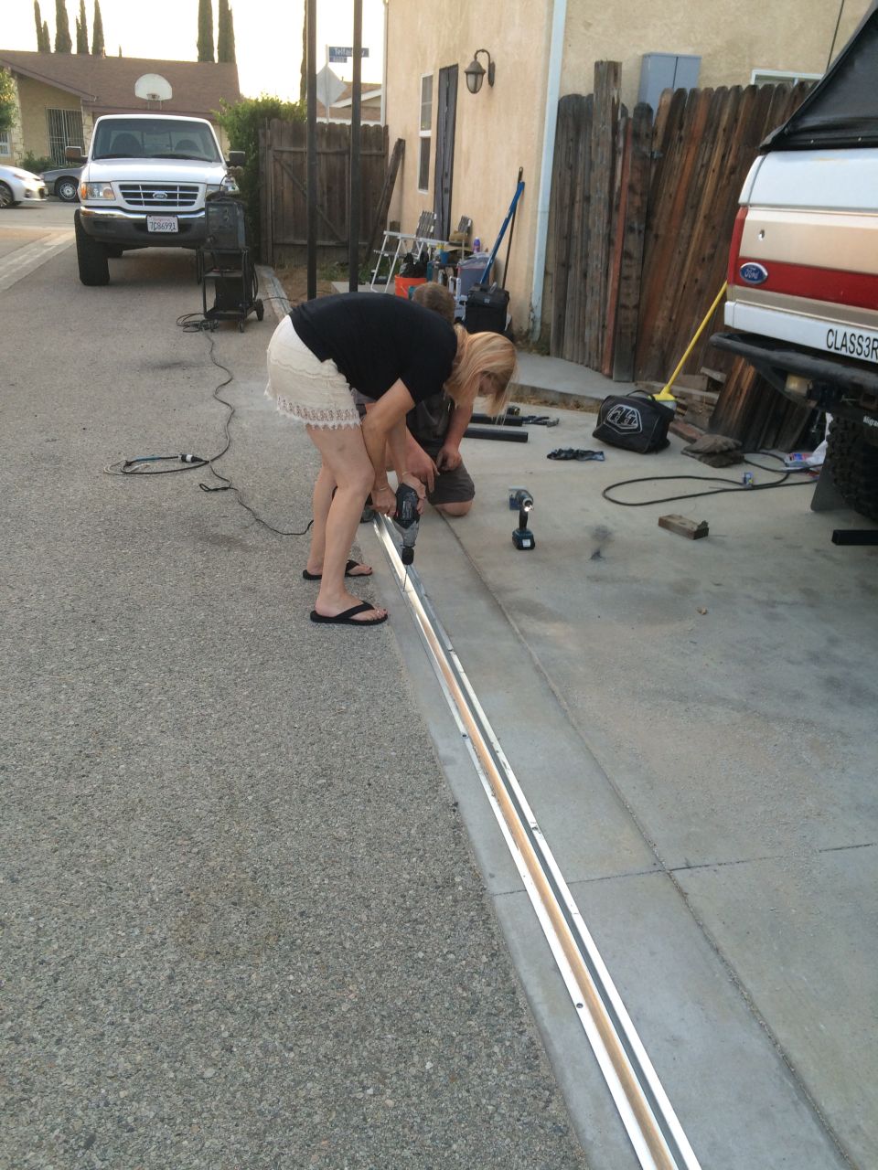 Two people work on a metal track installed in a driveway, with a white Ford pickup truck parked in the background