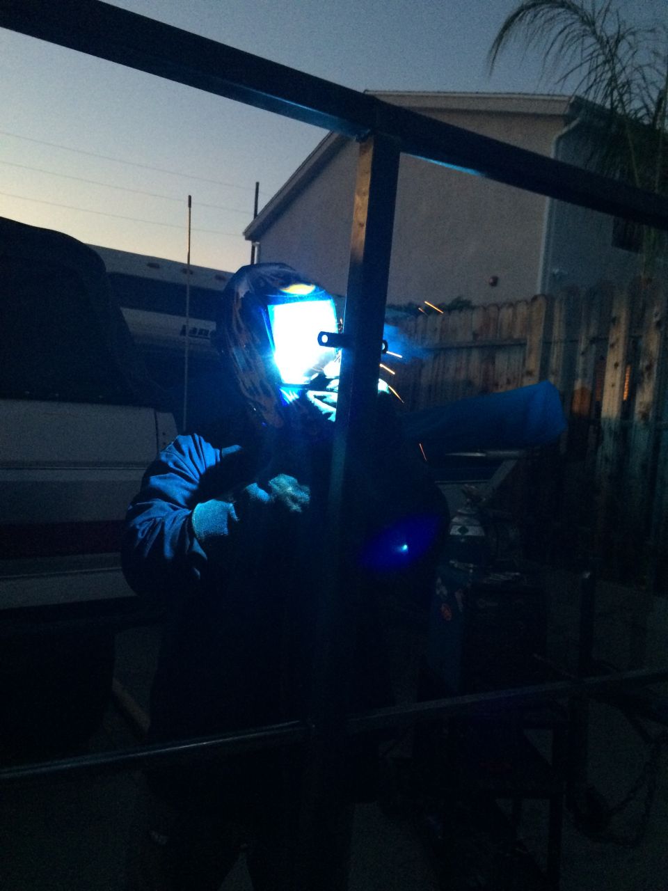 Person wearing a welding mask and protective gear, welding metal outdoors at dusk
