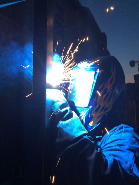 1968 Ford Mustang - A welder, illuminated by bright sparks and blue light, works on a metal structure against a darkening sky.