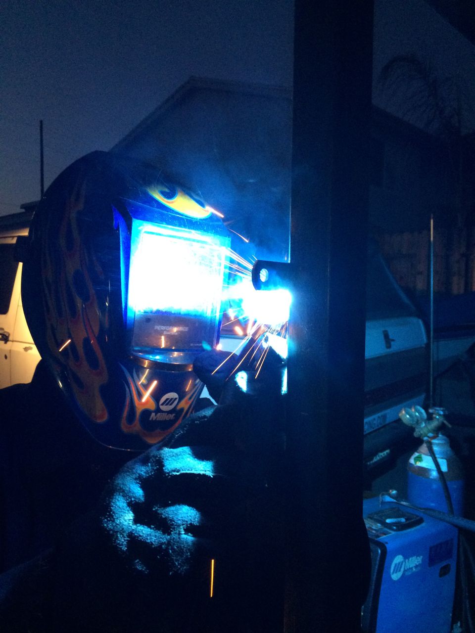 Welder wearing a Miller helmet with flames, sparks flying, working on metal in front of a dark building and a white vehicle