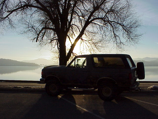 1989 Ford Bronco parked by a lake with a large tree and sunset in the background
