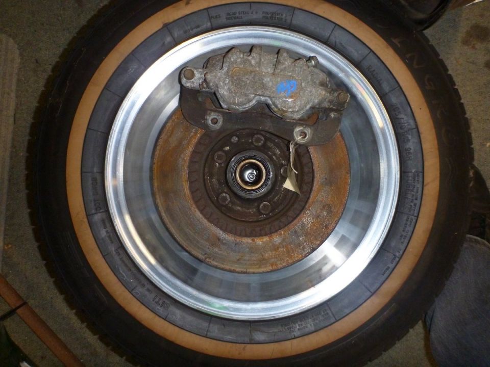Car brake rotor and caliper assembly with a tire in the background. Some text is visible on the tire sidewall