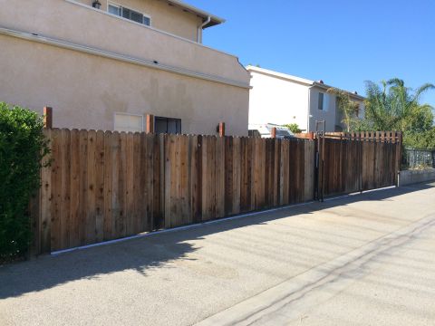 1968 Ford Mustang - A wooden fence and gate with a scalloped top edge stands beside a paved driveway, with houses and palm trees in the background under a clea…