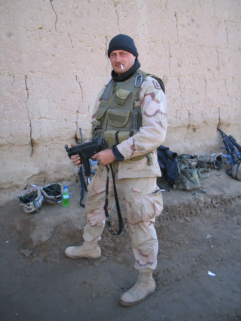 Soldier in desert camouflage uniform holding a submachine gun, standing in front of a mud-brick wall