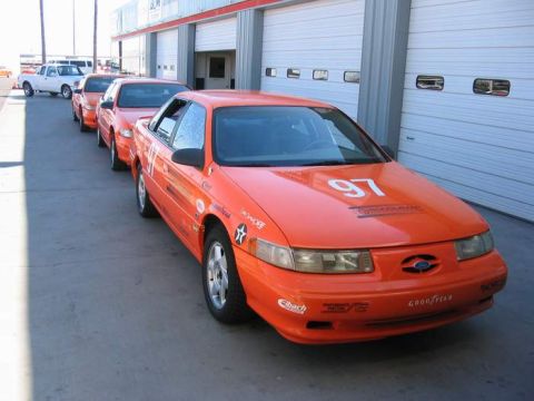 1993 Ford Taurus - A line of orange Ford Taurus race cars are parked outside a building with multiple garage doors.