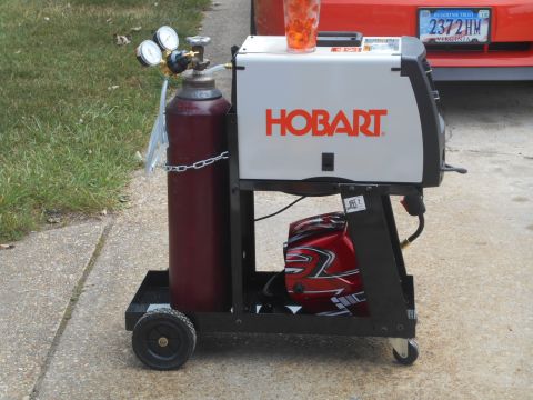 1994 Ford Bronco - Welding cart setup with gas cylinder and helmet, parked near a red car.