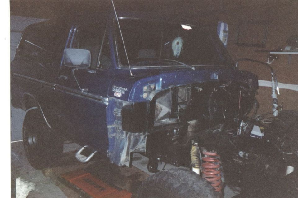 Blue Ford Bronco in garage with front end disassembled, exposing engine bay and red coil spring suspension