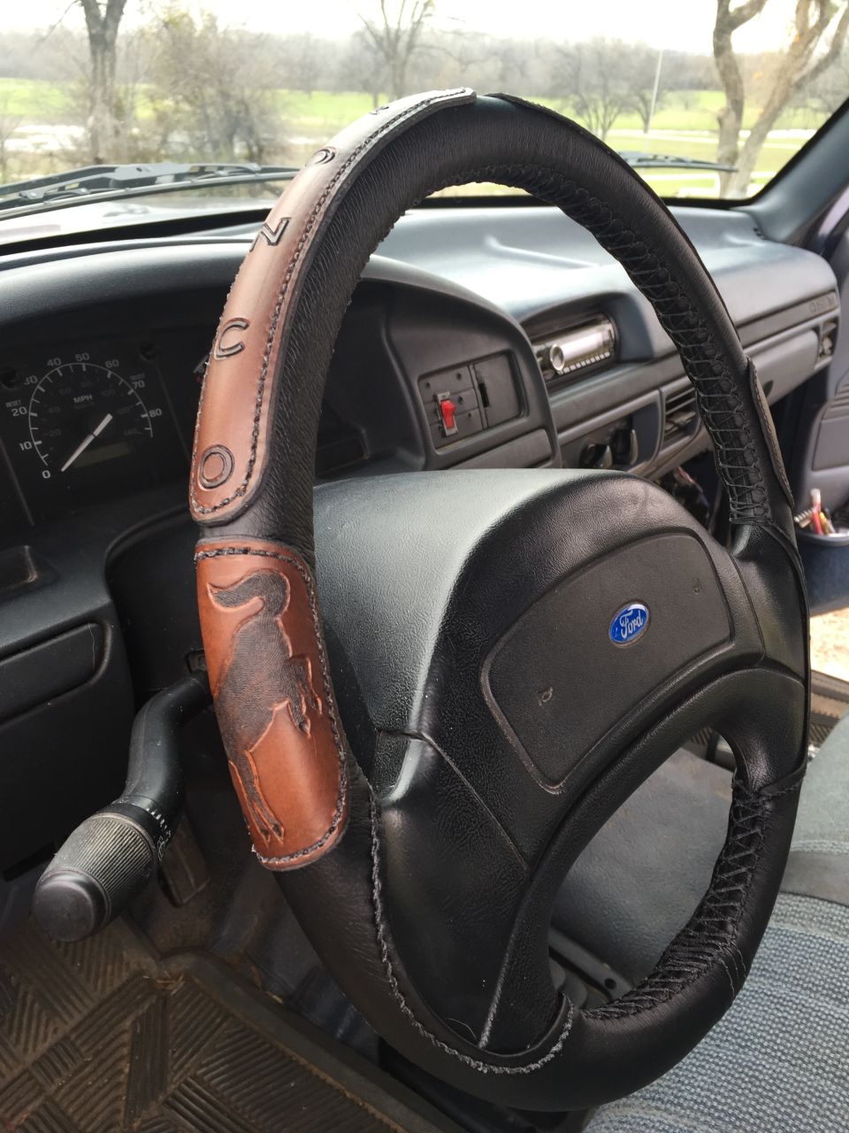 Interior shot of a 1993 Ford Bronco showing the steering wheel with a custom leather cover a Bronco emblem and the word 'BRONCO' stitched on it