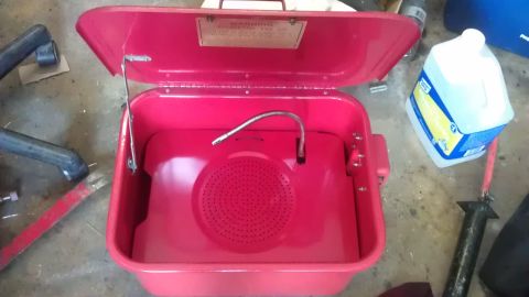 1994 Ford Bronco - A red parts washer with its lid open is shown in a garage setting. A jug of blue liquid is visible to the right of the parts washer.