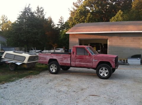 1991 Jeep Comanche - A red pickup truck is parked next to a garage, towing a camper shell on a trailer.