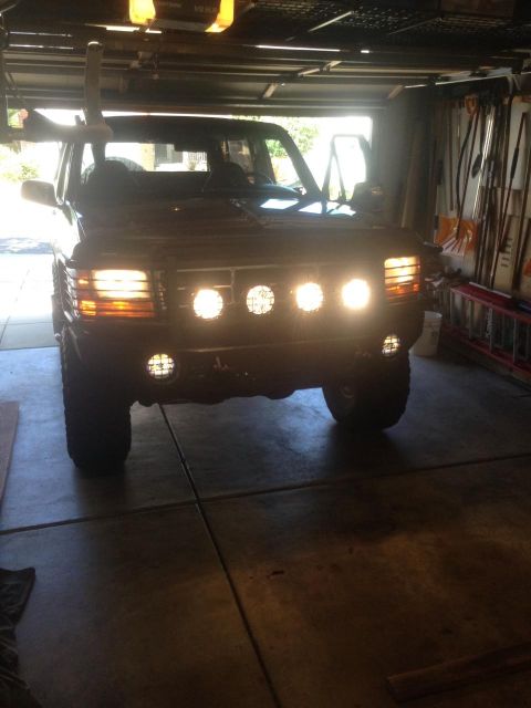 1996 Ford Bronco - Head-on view of a lifted full-size Ford SUV backlit in a garage doorway with its auxiliary lighting switched on: four round driving lights…