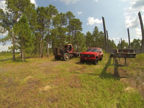 1995 Ford Bronco - A red 1990s Ford Bronco is parked in a grassy field next to a logging trailer and a red logging truck. The scene is set in a wooded area wi…