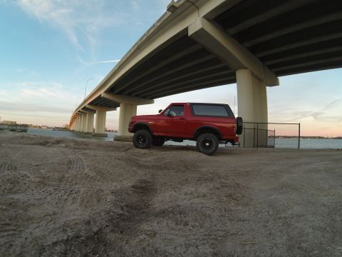 1995 Ford Bronco - A red 1990s Ford Bronco is parked on a sandy area under a large concrete bridge. The vehicle is viewed from the side, with the water and a…