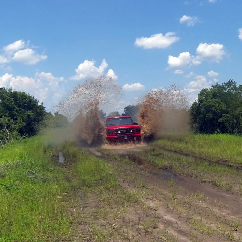 1995 Ford Bronco - A red Ford Bronco drives through a muddy track, kicking up large plumes of water and dirt on either side. The vehicle is seen head-on, with…