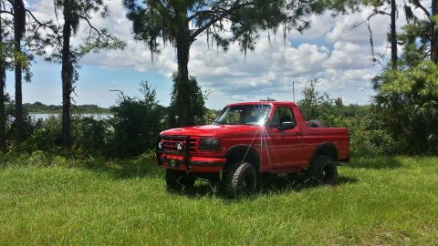 1995 Ford Bronco - A bright red, lifted Ford Bronco is parked in a grassy field with trees and a lake in the background under a partly cloudy sky.