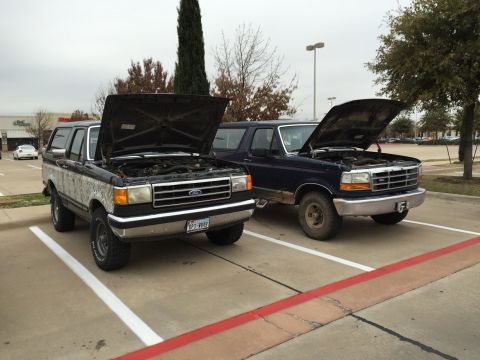 1993 Ford Bronco - Two Ford Bronco SUVs are parked side-by-side in a parking lot with their hoods open. The Bronco on the left is black with a camouflage patt…