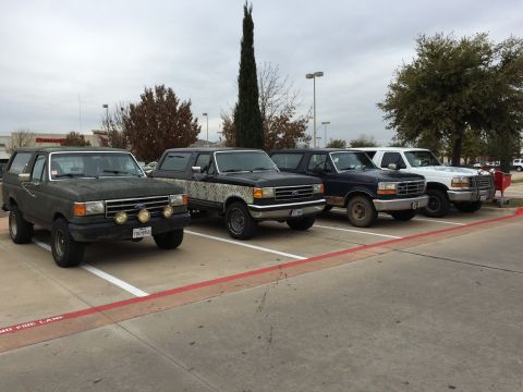 1993 Ford Bronco - Four Ford Bronco SUVs are parked in a row in a parking lot under an overcast sky. The vehicles are dark green, black with camouflage, dark…