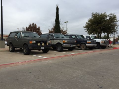 1993 Ford Bronco - A line of four Ford trucks and SUVs are parked in a parking lot under an overcast sky. The leftmost vehicle is a dark green Ford Bronco, fo…