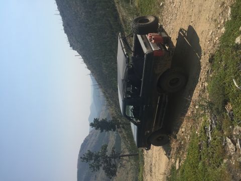 1994 Ford Bronco - A dark colored Ford Bronco is parked on a dirt road on a mountainside, with a spare tire and a red gas can mounted on the rear. The vehicle…