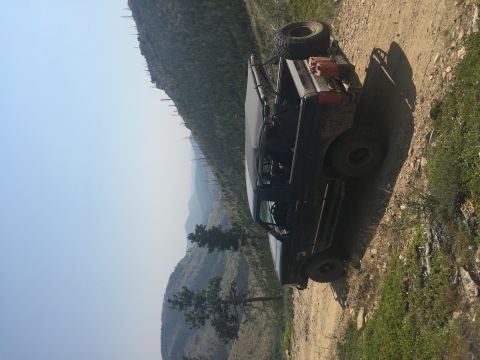 1994 Ford Bronco - A dark-colored Ford Bronco is parked on a dirt road on a mountainside with a spare tire mounted on the rear and a gas can on the roof rack.…
