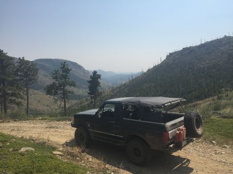 1994 Ford Bronco - A dark green Ford Bronco is parked on a dirt road overlooking a mountainous landscape under a clear blue sky. The Bronco has a soft top and…