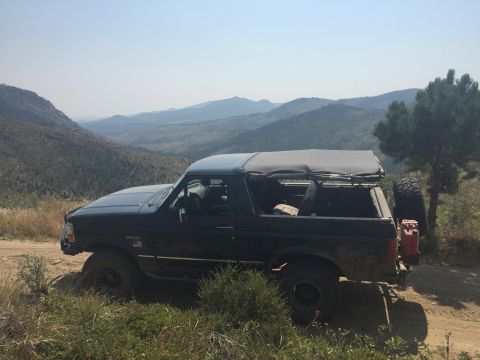1994 Ford Bronco - A dark-colored Ford Bronco with a soft top is parked on a dirt road overlooking a mountainous, tree-covered landscape. The vehicle has a sp…