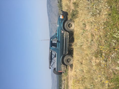 1994 Ford Bronco - A dark teal Ford Bronco with a soft top is parked on a grassy hillside under a clear blue sky. The vehicle is angled, showing its side prof…