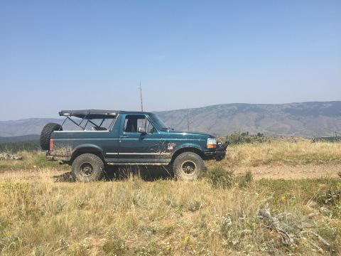 1994 Ford Bronco - A teal 1994 Ford Bronco is parked on a dirt road in a grassy, hilly landscape under a clear blue sky. The Bronco has a soft top, a spare ti…